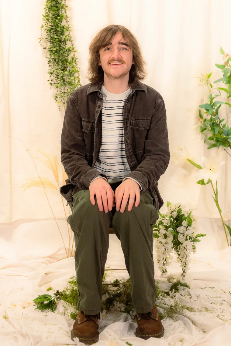 Drake Corrigan seated in a corduroy jacket and green trousers against a soft cream backdrop styled with trailing plants and white flowers.