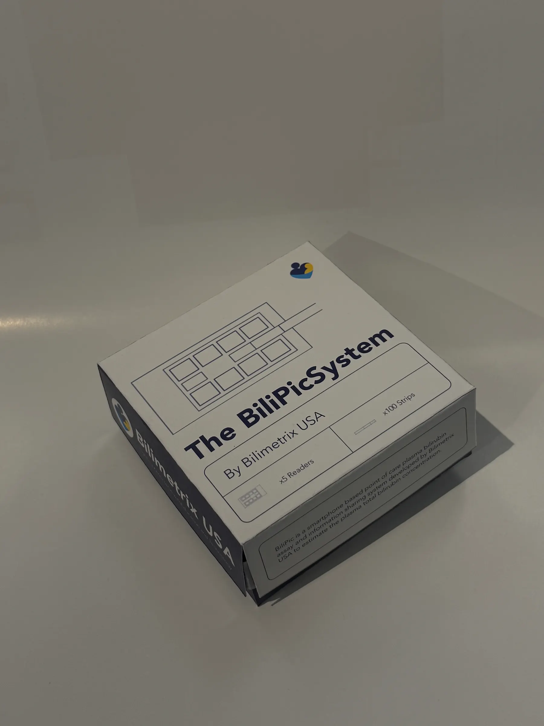 The printed package photographed closed on a white surface: white lid reading 'The BiliPic System / By Bilimetrix USA', a line diagram of the reader and strips, and 'x5 Readers / x100 Strips'.