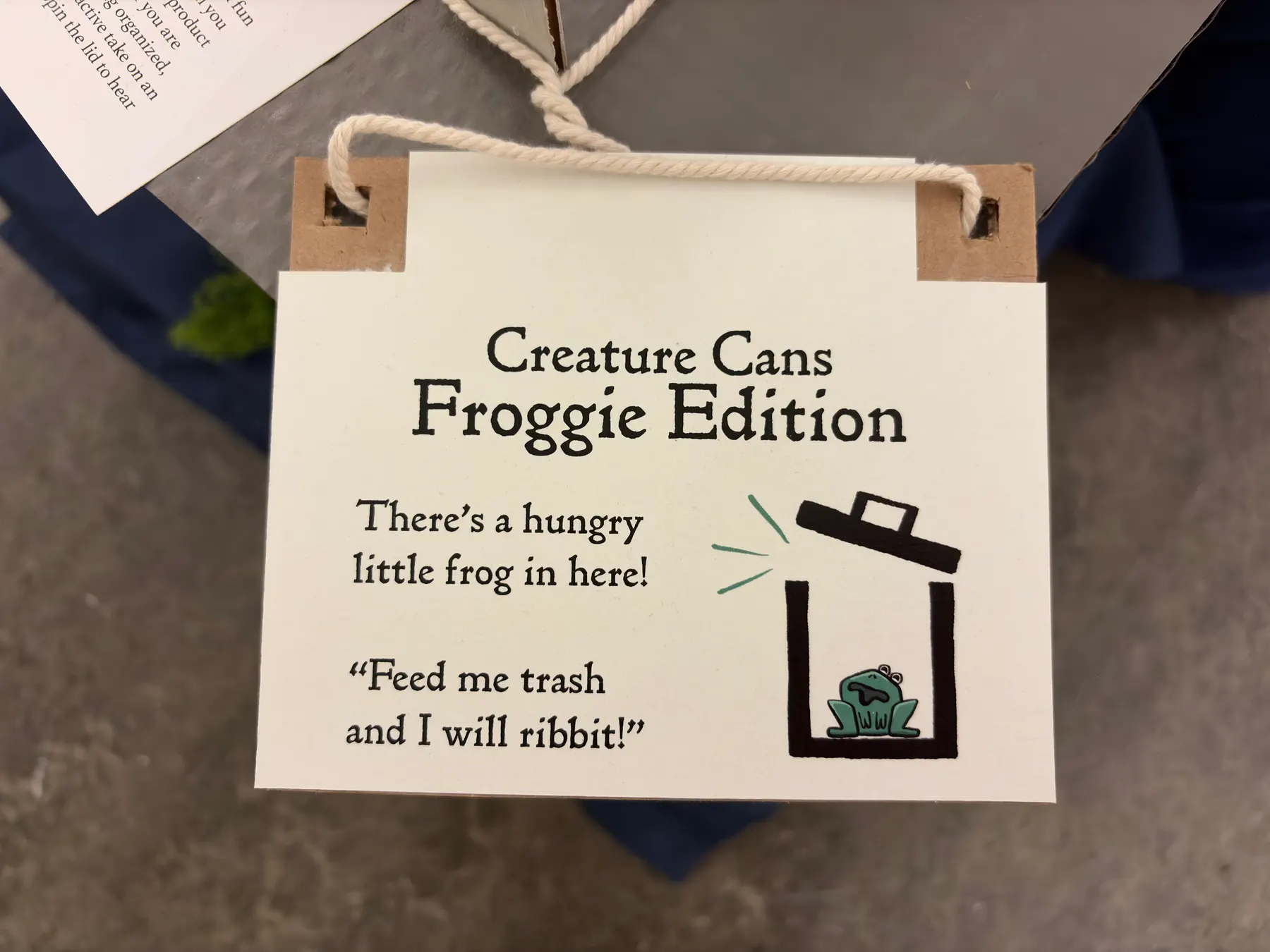 Detail of the Creature Cans cream hang tag with a serif logo and a small illustrated frog-in-a-bin icon, laced to the box with cord through cardboard eyelets.