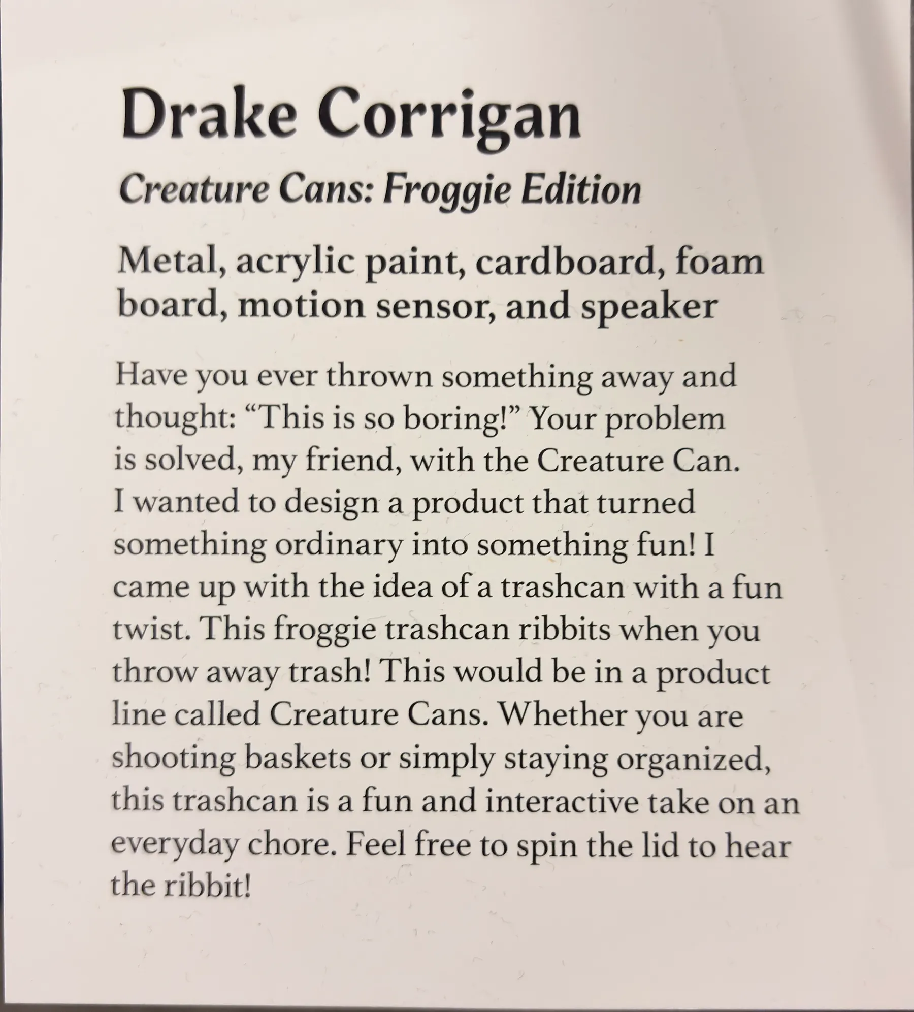 The printed exhibition placard for 'Creature Cans: Froggie Edition', listing the materials — metal, acrylic paint, cardboard, foam board, motion sensor and speaker — above a short description of the project.
